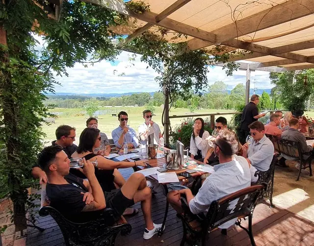 A large group at long tables under a leafy pergola