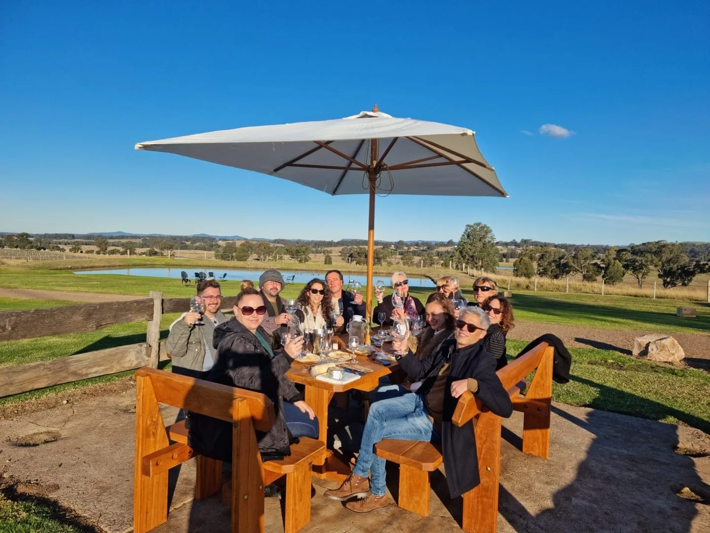 A tour group raising glasses around a long outdoor table at golden hour, Hunter Valley vineyards behind